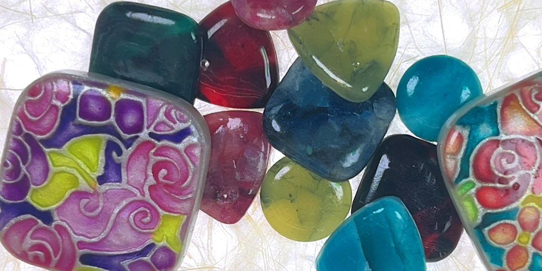 A close-up of colorful cabochons and on a textured white fabric background. The beads include smooth polished shapes in teal, green, red, and blue, and pink.