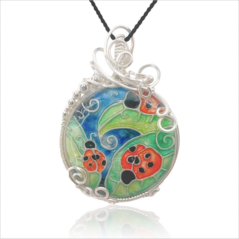 A pendant that looks like cloisonne, with illustrated ladybugs on leaves, in reds, greens and blues.